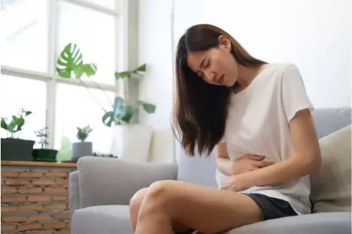 Woman with nausea holding stomach in pain sitting on her couch